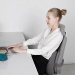 Femme assise devant un bureau sur une chaise avec un coussin ergonomique pour lombaire.
