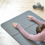 Tapis de yoga gris, pliable, sur lequel une femme en tenue de sport fait du yoga.