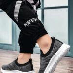 Femme portant des baskets orthopédique grises et noires, avec la semelle blanche. On ne voit que le bas de ses jambes et ses pieds.