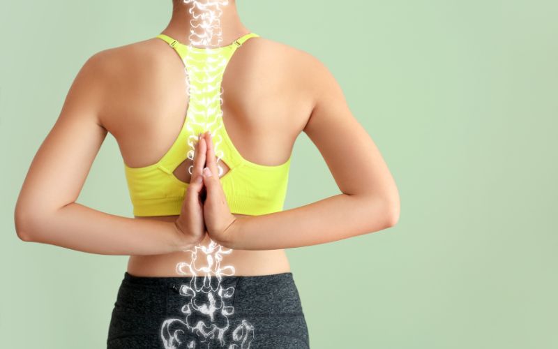 Femme de dos, en tenue de sport, joignant ses mains à plat au niveau de sa colonne vertébrale. En banc est dessiné les os de la colonne jusqu'au coccyx.