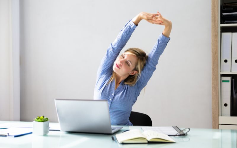 Femme assise devant un bureau, avec un ordinateur posé devant elle, elle s'étire le dos en levant les bras au-dessus de sa tête.