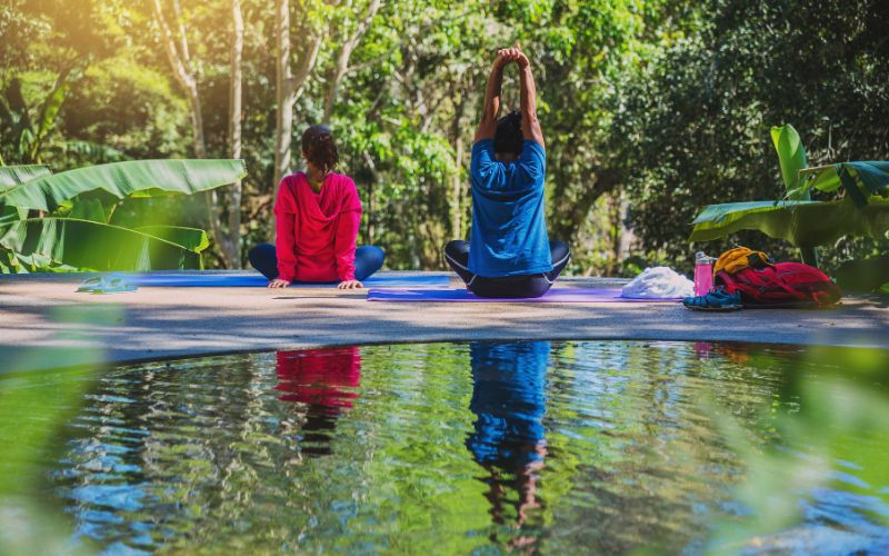 Une femme et un homme de dos assis en tailleur près d'un plan d'eau avec de la végétation autour d'eux, ils font des exercices d'étirements.