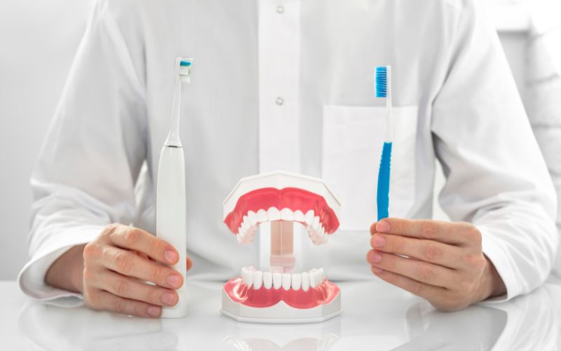 Personne en blouse blanche, devant une mâchoire de dentiste pour démonstration, dans une main, il tient une brosse à dent classique et dans l'autre une brosse à dents électrique.