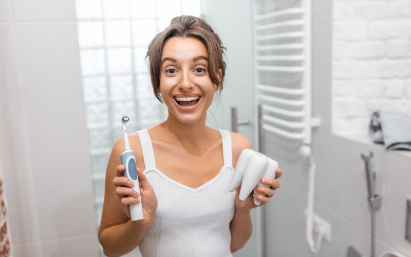 Femme dans sa salle de bain, avec une brosse à dents électrique dans une main et une dent de démonstration dans l'autre, elle fait un grand sourire.